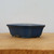 A rectangular blue glazed ceramic bonsai pot with a subtle matte finish, shown from a side profile resting on a light-colored natural wood surface. The pot features gently outward-sloping sides, a thick rounded rim, and four small integrated feet designed to provide elevation for drainage. Its deep blue color offers a clean, classic aesthetic for various bonsai species, set against a neutral, off-white background.