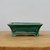 A side-profile view of a small, rectangular ceramic bonsai pot featuring a vibrant, glossy emerald green glaze. The pot has a slightly flared rim and decorative recessed panels on its sides. A fine, intricate network of "crazing" or crackle lines is visible throughout the glaze, providing a classic, artisanal texture. The vessel is elevated by four integrated corner feet with subtle arched cutouts between them for airflow and drainage. It sits centered on a light-colored, natural wood surface against a neutral, off-white background, highlighting the rich color and craftsmanship of the glazed ceramic.