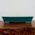 A horizontal studio shot showcases a rectangular, shallow-style bonsai pot featuring a rich, glossy teal-green glaze. The pot’s design includes a slightly protruding, rounded rim and four low, rectangular feet that create a graceful, subtle arch along its base. 

Close inspection reveals a fine, delicate crackle pattern throughout the deep green glaze, adding a sense of texture and traditional craftsmanship. The interior of the pot remains unglazed, showing a natural light tan ceramic color. The pot is positioned on a dark, polished wooden surface with a rustic, natural burl-like front edge, set against a plain, neutral off-white background. This elegant glazed ceramic pot is an example of the high-quality bonsai containers found at Bonsai Outlet.