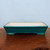 A rectangular, glazed bonsai pot in a deep emerald green or teal hue sits atop a dark, polished wooden surface. The shallow ceramic pot features slightly flared sides and a sturdy base with small, rounded feet at each corner. Its smooth, glossy exterior shows a faint crackle pattern in the glaze, while the interior is finished in a clean, off-white tone. The contrast between the rich green exterior and the light interior makes it a versatile choice for many bonsai species. The pot is positioned centrally against a simple, pale blue wall, highlighting its elegant shape and traditional design.