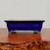 An eye-level, side-profile photograph features a rectangular, deep cobalt blue glazed ceramic bonsai pot. The pot has a glossy finish and a subtle indented rectangular panel on its front face. Its rim is slightly flared, while the four ornate feet at the corners feature intricate scrollwork carvings, where the natural cream-colored ceramic is visible beneath the blue glaze. The pot rests on a polished, reddish-brown wooden surface with a rich grain, and the foreground shows a rugged, natural-looking textured bark edge of the wood. The background is a clean, neutral, light-colored wall, which highlights the vibrant blue hue and elegant craftsmanship of the bonsai container.