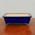 A deep rectangular glazed ceramic bonsai pot in a striking cobalt blue color sits centered on a polished, warm-toned wooden surface. The pot features a wide opening with a slightly flared rim and a subtle recessed panel running along its side. It is supported by elegant, cloud-shaped decorative feet at the corners, which show the natural, light-colored ceramic at their base. The interior of the pot is a contrasting pale cream or beige, typical of traditional ceramic craftsmanship. The pot is photographed from a low, straight-on angle against a plain, neutral light-grey wall, highlighting its clean lines, rich glossy finish, and classic design suitable for displaying a mature bonsai tree.