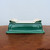 A high-quality, rectangular ceramic bonsai pot, model 2332b from Bonsai Outlet, is showcased on a polished, dark wood surface. The pot features a rich, deep emerald green glaze with a glossy finish and a subtle, intricate crackle pattern, often referred to as crazing. Its ornate design includes a stepped base and elegant, hand-finished scroll-like moldings at each of the four corners. The side profile shows indented rectangular panels that add depth and character to the piece. From this slightly elevated angle, the creamy, off-white interior of the pot is visible, revealing four essential drainage holes at the bottom. The pot is set against a plain, light-colored background, which emphasizes its vibrant color and sophisticated, traditional craftsmanship, making it an ideal choice for displaying a prized bonsai tree.