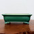 An emerald green rectangular glazed ceramic bonsai pot, shown from a side profile. The pot features a glossy finish with a fine network of crackle glaze, also known as crazing, which adds a vintage, textured appearance. Its design includes an indented rectangular panel on the front and sides for a tiered look, leading up to a slightly flared top rim. The pot is elevated on four ornate, cloud-scrolled feet that match the green glaze. It is resting on a dark, rustic wooden surface with a plain, off-white background, highlighting its deep color and decorative details.