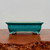 A rectangular turquoise glazed bonsai pot with a fine crackle finish, displayed on a rustic, dark wood surface against a plain, light gray background. This ceramic bonsai container features a vibrant teal-blue glaze with intricate crazing lines across its entire surface. The pot has a slightly flared top rim and raised decorative rectangular panels on its sides. It is supported by four ornate, scrolled feet that reveal the natural, unglazed cream-colored clay underneath at the tips. The interior of the pot also shows the natural tan clay color. The low-profile design and elegant glaze make it a traditional and sophisticated choice for various bonsai tree species.