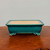 A rectangular glazed bonsai pot is shown in a front-facing, eye-level shot. The pot is finished in a vibrant, deep teal or turquoise glaze that features a delicate, all-over crackle pattern. It has rounded corners and a slightly flared rim, with a subtle rectangular recessed panel on its front face.

The pot sits on four ornate, scroll-shaped feet, often referred to as cloud feet, which reveal the natural light tan-colored ceramic underneath the teal glaze. The interior of the pot is unglazed and matches the light beige clay of the feet. The pot is centered on a dark, polished wood surface with a visible grain, set against a neutral, light-colored wall, emphasizing the rich color and craftsmanship of the bonsai container.