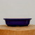 A detailed product shot of a shallow, deep blue glazed bonsai pot (product code blue-2337a) from Bonsai Outlet. The ceramic pot features a rich, glossy cobalt finish and an elegant mokko or lobed rectangular shape with soft, rounded corners and a slightly flared lip. It is positioned at eye-level on a thick, natural-grained wooden block against a plain, off-white background. The high-shine glaze reflects subtle light, while the unglazed bottom edge and small, carved feet are visible, highlighting its functional design for drainage. The interior of the pot is a pale tan ceramic color. This vibrant blue glazed container is a classic choice for showcasing deciduous or flowering bonsai trees, providing a striking color contrast to green foliage.