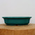 A high-angle, eye-level photograph showcases a rectangular, teal-green glazed bonsai pot resting on a thick, natural wood slab. The pot features a rich, glossy finish with a fine crackle pattern throughout its deep turquoise surface. It is designed with a prominent, slightly flared rim and elegant lobed corners, giving it a soft, rounded rectangular shape. Along the very top edge of the rim and the bottom feet, the pale, unglazed buff-colored ceramic is visible, providing a subtle contrast to the vibrant green glaze. The pot sits on a light brown wooden base with a visible natural grain, all set against a clean, neutral off-white background that emphasizes the pot's color and form. Small cutouts at the base serve as integrated feet, essential for drainage in bonsai cultivation.