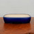 A front-facing, eye-level photograph captures a rectangular, glazed ceramic bonsai pot resting on a rustic wooden surface. The pot’s exterior is finished in a vibrant, glossy cobalt blue glaze that catches the light, while the interior is a smooth, contrasting cream or off-white color. Its design features soft, rounded corners and gently bowed sides, giving it an elegant, classic silhouette. The pot sits on a slightly recessed base with four small, integrated feet, a common feature in functional bonsai containers to allow for proper drainage. The dark, textured wood grain of the tabletop highlights the pot's deep blue hue, while the clean, neutral off-white background keeps the focus entirely on the craftsmanship and color of the glazed bonsai pot.