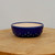 A detailed view of a small, round ceramic bonsai pot featuring a vibrant, glossy cobalt blue glaze. The shallow pot is decorated with a series of raised, circular studs or bosses along the bottom half, adding a textured, artisanal feel. Its smooth rim encloses an unglazed, light beige interior, typical of functional bonsai containers. The pot sits centered on a thick, natural wood slab with visible grain patterns, set against a plain, neutral off-white background that makes the rich blue color pop. This elegant piece represents a classic style of glazed bonsai pots used for accenting miniature trees.