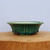 A side profile of a shallow, mokhoke-style glazed bonsai pot in a rich forest green color. The pot features a wide, flared rim with decorative scalloped edges and vertical ribs that flow down to small, integrated feet. The glossy green glaze has a fine crackle pattern, while the interior is a contrasting light cream ceramic. The pot sits on a flat-topped, light-colored wooden log or block with visible wood grain, positioned against a plain, off-white background.