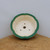 An overhead view of a handcrafted round ceramic bonsai pot with a distinctive scalloped, flower-shaped rim. The rim is finished in a vibrant, deep emerald green glaze that contrasts beautifully with the pot’s smooth, creamy off-white interior. Visible at the base are several functional features, including one large central drainage hole and three smaller holes used for wiring a bonsai tree securely in place. The pot is presented on a rustic, light-toned wooden surface with a visible grain, set against a plain, neutral background. This elegant green glazed bonsai pot from Bonsai Outlet is designed to provide both aesthetic appeal and the proper drainage and support needed for healthy bonsai growth.
