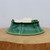 An emerald green glazed ceramic bonsai pot is shown from a side-view perspective while positioned upside down on a thick, light-colored wooden block. The pot features a rich, deep green glaze with a fine, intricate crackle pattern, often referred to as crazing, which adds a decorative, antique-like texture to the surface. Its elegant, flaring shape is accented by subtle vertical ridges that divide the body into lobes, ending in a wide, scalloped rim that resembles the petals of a flower. The unglazed white ceramic underside is visible, showcasing four sturdy, rectangular feet and several pre-drilled holes essential for bonsai care: one large central drainage hole and multiple smaller holes intended for anchoring bonsai wire. The pot rests on a natural wood grain surface against a minimalist, neutral light gray background, highlighting its vibrant color and artisanal craftsmanship.