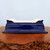 An eye-level, side-view photograph of a rectangular, blue glazed bonsai pot from Bonsai Outlet. The pot features a rich, deep cobalt blue glaze with a highly reflective, glossy finish that catches the light. Its design is characterized by slightly flared sides and a prominent, flat lip at the base. At the top four corners, there are small, decorative scroll-like accents in the same blue glaze. The interior of the pot, visible from the top edge, is a contrasting unglazed or off-white ceramic. The pot is showcased on a thick, irregularly shaped natural wood burl slab with a warm, reddish-brown tone and intricate organic texture. The entire scene is set against a neutral, light-colored background, emphasizing the elegant color and form of the ceramic bonsai container.