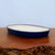 A shallow and sleek oval glazed bonsai pot with a vibrant cobalt blue exterior and a smooth, off-white interior. The low-profile ceramic container sits on a rustic, dark-toned wooden table with a live-edge texture. The interior of the pot shows small drainage holes, making it ideal for the health of a bonsai tree. The glossy blue finish provides a bold contrast against the natural wood grain and the neutral, light-colored backdrop, showcasing a classic and elegant vessel for bonsai cultivation.