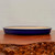 A side-view, eye-level photograph of a shallow, oval-shaped ceramic bonsai pot. The pot features a vibrant, deep cobalt blue glaze on its exterior walls and along its slightly rounded rim. Its interior remains unglazed, showing a smooth, light tan natural clay surface. The low-profile pot rests on a polished, dark reddish-brown wooden display table, which has a distinct, rugged burl wood texture visible in the foreground. The entire scene is set against a minimalist, neutral light-gray background that emphasizes the rich blue color and clean lines of the glazed bonsai pot.