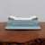 A side view of a rectangular, light blue glazed ceramic bonsai pot resting on a weathered, dark wood surface. The pot has a low profile with a slightly flared base and decorative, white cloud-style scrollwork at each of its four corners. The exterior is finished in a smooth, pale blue glaze, while the top surface is flat and white, featuring several small circular holes intended for drainage and wiring a bonsai tree. The background is a simple, solid off-white wall, emphasizing the pot's clean lines and elegant design. This high-quality glazed bonsai pot combines functional features with a classic, decorative aesthetic suitable for various tree styles.