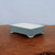 A rectangular, light blue glazed ceramic bonsai pot sits centered on a polished, rich brown wooden surface with a visible natural grain. The pot features a soft, matte powder-blue glaze on its exterior walls and thin top rim, while the interior is a contrasting creamy off-white color. It has a shallow profile and is supported by four small, outward-curving cloud-style feet. The entire scene is set against a plain, neutral light grey background, emphasizing the clean lines and delicate color of the pot.