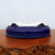 A close-up, slightly high-angle shot shows a shallow, oval-shaped ceramic bonsai pot resting upside down on a rustic, live-edge wooden stand. The pot is finished in a rich, high-gloss cobalt blue glaze that reflects the surrounding light. Around the lower perimeter of the pot is a decorative "drum-pot" style border, characterized by a row of small, raised circular beads or rivets. 

The pot features four sturdy, integrated feet glazed in the same deep blue. Because it is positioned upside down, the interior and base of the pot are visible, revealing a light-colored, unglazed clay surface with pre-drilled drainage holes essential for maintaining the health of a bonsai tree. The dark, textured wood of the stand provides a natural contrast to the smooth, polished finish of the blue glazed pot, all set against a clean, neutral-toned background.