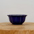 A small, round cobalt blue glazed ceramic bonsai pot with a scalloped, lobed exterior design that tapers toward a footed base. The deep blue glossy glaze features delicate, lighter highlights along the vertical ridges and the flared rim, creating a textured, petal-like appearance. The pot is positioned in the center of the frame, resting on a rustic, light-toned wooden cross-section with visible natural grain. The background is a clean, neutral off-white, making the vibrant blue of the bonsai container stand out as the primary focus.