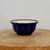 A small, round ceramic bonsai pot with a deep cobalt blue glaze and a decorative fluted design. The pot features vertical ridges and a scalloped rim, with the glaze appearing slightly lighter on the edges of the ridges, highlighting the pot's texture. The interior of the pot is a contrasting unglazed cream color. It is displayed on a thick, rustic wooden round with visible grain patterns and a natural crack, all set against a clean, minimalist off-white background.