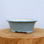 A side-view studio photograph shows a small, light blue glazed ceramic bonsai pot resting on a thick, natural wood block. This shallow, oval-shaped container features a classic scalloped "mokkou" or four-lobed design with a wide, flared rim. The exterior is finished in a smooth, uniform pale blue glaze that reflects light softly, while the interior shows the natural, off-white tone of the ceramic clay. The pot is elevated by four integrated, decorative feet with a gentle wave pattern. Set against a plain, neutral white background, the image highlights the elegant form and calming color of this bonsai container, making it an ideal choice for various bonsai tree styles.