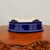 An ornate, deep cobalt blue glazed ceramic bonsai pot is shown from a low angle, highlighting its intricate craftsmanship. The pot is positioned on its side or slightly overturned to showcase its decorative, cloud-shaped feet, which feature elegant scrollwork details. These feet, along with the very bottom of the pot, are left unglazed, revealing a smooth, off-white ceramic material that contrasts beautifully with the rich blue exterior.

The sides of the pot are covered in a brilliant, glossy blue glaze that reflects light, emphasizing its smooth, rounded-rectangular or oval shape and slightly flared rim. This high-quality bonsai pot is displayed on a rustic, polished wood slab with a natural, textured burl edge, providing a warm, organic contrast to the cool tones of the ceramic. The entire scene is set against a neutral, off-white background, making the vibrant blue of the pot the central focus. This image highlights the aesthetic appeal and functional design of a classic glazed bonsai pot from Bonsai Outlet.