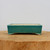 A rectangular, glazed ceramic bonsai pot in a vibrant seafoam or teal green color is centered on a light-colored wooden pedestal. This bonsai container features a smooth, glossy finish with a subtle crackle glaze pattern visible upon closer inspection. The pot's design includes slightly rounded corners, a flat top rim, and four small, flared feet that elevate the base. The interior of the pot and the bottom of its feet reveal a pale, unglazed cream-colored clay. Set against a plain, neutral background, the pot's rich green hue stands out, making it an ideal choice for various bonsai tree species.