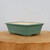 A side-view of a rectangular matte green glazed bonsai pot resting on a thick, natural wood slice. The ceramic pot features a slightly flared rim, tapered sides, and a subtle decorative ridge near its base, which is supported by four small integrated feet. The glaze has a soft, earthy teal-green finish, while the interior of the pot shows a pale, unglazed ceramic surface. The composition is clean and minimalist, set against a plain light gray background to highlight the pot's functional design for bonsai cultivation.