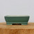A side-view photo of a rectangular ceramic bonsai pot finished in a soft, matte seafoam green glaze. The pot has a classic, clean design with slightly rounded corners and sides that taper gently toward the base. A decorative raised line runs horizontally near the bottom, just above four integrated, sturdy feet. The cream-colored ceramic body of the pot subtly peeks through the glaze along the rim and at the base of the feet, adding a sense of artisanal character. The pot is presented on a thick, natural wood block that highlights its grain and texture, all set against a neutral, off-white background. This professional-grade glazed bonsai pot combines elegance with functionality, making it a versatile choice for a variety of bonsai tree styles and species.