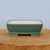 A detailed view of a rectangular green glazed bonsai pot with a smooth, matte sage-green finish and softly rounded corners. The pot is positioned on a light-colored, natural wood platter, which shows the grain and texture of the wood. A subtle decorative molding runs along the lower portion of the pot, just above its sturdy, integrated rectangular feet. The interior of the ceramic bonsai container is a contrasting off-white or cream color. The entire scene is set against a clean, neutral light blue-grey background, emphasizing the pot's classic aesthetic and suitability for housing a miniature tree.