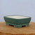 A rectangular glazed ceramic bonsai pot in a muted teal-green color, viewed from a three-quarter angle. The pot has gently rounded corners, inward-sloping sides, and a subtle rim detail around its base. It stands on four integrated, blocky feet that show the natural, off-white color of the unglazed ceramic on their bottom surfaces. The interior of the pot is also unglazed off-white. This professional-grade bonsai container is set upon a rustic, light-brown wooden tabletop against a clean, pale blue background, highlighting its elegant design and earthy green finish.