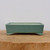A shallow, rectangular ceramic bonsai pot finished in a smooth, matte seafoam green glaze sits centered on a rustic wooden base. The pot features a clean, minimalist design with four small, decorative feet at each corner that flare slightly outward, providing a stable and elegant lift. The interior edge of the pot reveals a contrasting unglazed, cream-colored ceramic body. It is positioned on a thick, light-brown wooden pedestal with a prominent natural grain and rough-hewn edges, suggesting a cross-section of a tree trunk. The entire setup is photographed against a neutral, off-white background with soft, even lighting that highlights the subtle texture of the green glaze and the organic feel of the wood, presenting a professional and serene product display for a glazed bonsai pot.