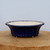 A side-view studio photograph showcases a shallow, deep cobalt blue glazed ceramic bonsai pot. The pot features a distinct scalloped or lobed rim, giving it a floral, lotus-like appearance. The rich blue glaze has a glossy finish that reflects light, while the interior of the pot is a contrasting, unglazed off-white or cream color. 

The pot is designed with a flared shape and sits on several small, integrated feet that lift it slightly off the surface for drainage. It is positioned in the center of a thick, natural wood slice with visible grain and a light tan hue. The background is a clean, minimalist pale blue-grey wall, creating a simple and elegant composition that highlights the craftsmanship and vibrant color of the glazed bonsai pot.