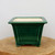 A deep green glazed ceramic bonsai pot featuring a classic square shape with a tapering profile and a flared top rim. The glossy forest green glaze has a subtle crackled texture and covers a decorative raised rectangular panel on the front face. This deep bonsai pot sits on four small, ornate feet on a rustic wooden display slab against a neutral off-white background. The interior of the pot remains unglazed in a natural cream-colored ceramic.