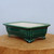 An emerald green glazed ceramic bonsai pot in a rectangular shape sits on a rustic wooden block against a neutral light grey background. This traditional container features a glossy, deep green finish with a subtle crackle glaze and decorative recessed panels on each side. The pot is elevated by four ornate scrolled feet, often referred to as "cloud feet," and has a contrasting cream-colored interior. The wide rim and elegant design make it an ideal choice for housing a variety of bonsai trees.