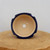 A top-down view of a small, glazed ceramic bonsai pot in a rich dark blue finish. The pot is positioned on a thick, natural wood block against a plain, off-white background. It features a decorative scalloped rim with four symmetrical notches, giving it a unique geometric silhouette. The exterior and rim are finished with a glossy cobalt blue glaze, while the interior remains unglazed, revealing the natural beige-colored clay. The bottom of the pot contains five holes: one large central drainage hole and four smaller surrounding holes intended for wiring a bonsai tree securely in place.