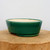A side-profile photograph of an oval, glazed ceramic bonsai pot in a deep, glossy forest green color. The pot's exterior features a smooth, reflective finish with slightly sloping walls and a subtle decorative ridge around the base. It is elevated by small, unglazed feet for drainage. The interior of the shallow pot is unglazed, revealing a light cream-colored ceramic body. The container is centered on a thick, rustic wooden pedestal made from a slice of a tree trunk, set against a plain, off-white background.
