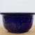 An eye-level, side-view image of a deep cobalt blue glazed ceramic bonsai pot, style number 2085. The pot is round and shallow with a classic, wide, flared rim that tapers down to a smaller base. Its exterior features a hand-modeled, textured surface with rippled indentations, creating a play of highlights and shadows across the rich, glossy finish. The vibrant blue glaze has a high-shine quality, reflecting its surroundings. The pot is presented on a light-colored wooden surface against a plain, off-white background, emphasizing its bold color and artistic detail. This type of glazed bonsai container is an excellent choice for a variety of tree species, providing a striking contrast to green leaves or bright seasonal foliage.