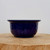 A side-view studio photograph of a deep cobalt blue glazed bonsai pot resting on a circular, natural wood slice. The ceramic pot features a rounded body with a subtly hammered or dimpled exterior texture that creates variations in light and shadow across its high-gloss finish. The pot has a wide, flared rim at the top and tapers slightly down to a small, footed base. The deep navy blue color is vibrant and glossy, reflecting soft light that showcases the textured surface. The background is a clean, minimalist off-white, highlighting the craftsmanship and rich color of this traditional bonsai container.