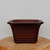 A deep reddish-brown unglazed ceramic bonsai pot is centered on a polished natural wood slice against a plain, light-gray background. The pot features a traditional rectangular shape with elegant clipped corners, giving it a subtle octagonal profile. It has a flared top rim and a decorative recessed rectangular panel on its front face, adding a touch of texture to its smooth, matte finish. The pot is supported by a tiered base with four short, sturdy block feet. This classic, earth-toned bonsai container is designed with a timeless aesthetic suitable for a variety of bonsai tree styles.