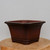 A high-quality, unglazed bonsai pot made of dark reddish-brown ceramic clay, positioned centrally on a light-colored wooden pedestal. This square-shaped pot with chamfered corners creates an octagonal silhouette, featuring a wide, outward-flaring rim and decorative recessed panels on each side. The matte finish and earthy tones of the unglazed clay provide a classic, natural look suitable for various bonsai styles. At its base, the container is supported by four small, sturdy feet, ensuring proper drainage and airflow. The simple, neutral gray background emphasizes the pot's elegant lines and solid construction, making it an ideal choice for a sophisticated bonsai display.