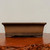 A rectangular unglazed bonsai pot in a deep, earthy chocolate-brown clay, shown from a front-facing eye-level perspective. The pot features a wide, flared upper rim and sides that taper slightly inward toward a decorative, double-banded base. It is supported by small, sturdy rectangular feet at each corner. The unglazed finish gives it a natural, matte texture with a fine-grained appearance. The pot sits on a polished, dark reddish-brown wooden table with a rugged, natural-edge front. The background is a plain, light-colored wall, focusing all attention on the clean lines and traditional craftsmanship of the ceramic bonsai container.