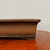 A close-up, side profile view of a rectangular unglazed bonsai pot, highlighting its fine-grained, earthy brown clay texture. The pot's design features elegant horizontal molding lines near the top rim and bottom base, giving it a classic and sturdy appearance. A single, short corner foot is visible, designed for proper drainage and airflow. The pot is showcased on a polished, reddish-brown wooden surface against a plain, off-white backdrop, emphasizing the natural beauty and artisanal quality of traditional unglazed bonsai containers.