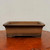 A classic rectangular unglazed bonsai pot crafted from earthy brown clay, featuring a matte finish and a wide, outward-flaring rim. The container sits on a polished reddish-brown wooden display slab against a clean, off-white backdrop. Its design includes a subtle horizontal recessed line along the upper body and a sturdy base with distinct feet at each corner. This traditional-style pot provides a neutral, natural aesthetic ideal for housing coniferous or deciduous bonsai tree species.