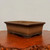 A rectangular unglazed bonsai pot sits at a slight angle on a polished, dark reddish-brown wood table. The ceramic pot is a warm, medium brown with a smooth, matte texture and subtle color variations typical of high-quality clay. It features a prominent, outward-flared rim at the top and a decorative, stepped base. Four short, blocky feet at the corners elevate the container, providing space for drainage and air circulation. The table underneath has a rustic, live-edge border with a deep, glossy wood grain that reflects the light. Behind the pot, a plain, off-white wall provides a simple, neutral backdrop, emphasizing the clean lines and earthy tones of this traditional unglazed bonsai pot.