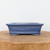 A rectangular, seven-inch blue glazed ceramic bonsai pot sits centered on a rustic, natural wood surface against a minimalist white background. The bonsai container features a wide, flared lip at the top and a subtle decorative ridge encircling the base, which rests on four small, integrated feet. The glaze is a vibrant, slightly mottled shade of medium blue, providing a classic and elegant aesthetic. The smooth, glazed finish and traditional shape make it an ideal choice for showcasing various bonsai tree styles. The soft, even lighting in the product photograph emphasizes the pot's texture and rich blue color.