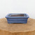 A seven-inch rectangular glazed ceramic bonsai pot in a vibrant, mottled blue hue is centered in this product shot. The pot features a classic design with a slightly flared, stepped rim and a matching stepped base that rests on four integrated feet. The blue glaze exhibits a subtle, speckled texture, with some lighter pale blue or white tones appearing like frosting around the upper interior edge. The inside of the pot is unglazed, showing the dark, reddish-brown clay body. The bonsai pot sits atop a thick, rustic slab of natural wood with a visible grain and soft texture. The entire composition is set against a plain, solid white background, creating a clean and minimalist aesthetic that draws all attention to the details and craftsmanship of the blue glazed bonsai pot.