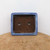 A top-down view of a rectangular seven-inch blue glazed bonsai pot. The pot features a distinctive speckled blue glaze on its wide, flat upper rim. The interior is unglazed, revealing the natural dark brown terracotta-colored clay. Symmetrically placed at the bottom are two large circular drainage holes and four smaller holes designed for securing bonsai wire. The pot is showcased on a weathered, light brown wooden surface against a clean white wall, highlighting its color and craftsmanship.