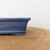 A detailed close-up profile view captures the side and corner of a seven-inch rectangular glazed blue bonsai pot. The ceramic pot is finished in a rich, deep blue glaze with fine white speckles, giving it a textured appearance. Its design includes a subtly flared upper rim and a distinct horizontal decorative line encircling the base, just above the low, integrated feet. The pot sits on a flat, light brown wooden surface, with a plain, off-white wall serving as a clean and simple backdrop, emphasizing the pot's color and craftsmanship.